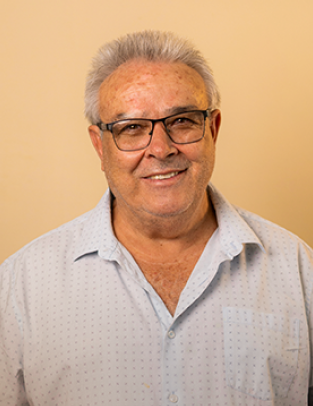 Tião Xavier sorrindo, de óculos e camisa clara, diante de uma parede bege